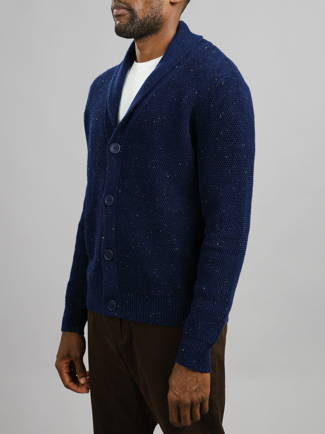 A man stands with arms by his sides in a blue Vermont Shawl Collar Cardigan from ID Menswear, made of speckled yarn and featuring buttons, over a white undershirt. He pairs it with brown pants against a plain gray background.