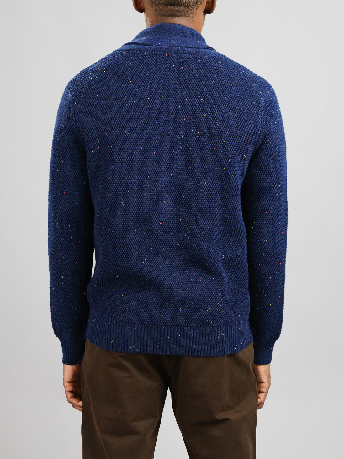 A person in a Vermont Shawl Collar Cardigan by ID Menswear, featuring a dark blue speckled pattern and paired with brown pants, is facing away from the camera against a plain light gray background.