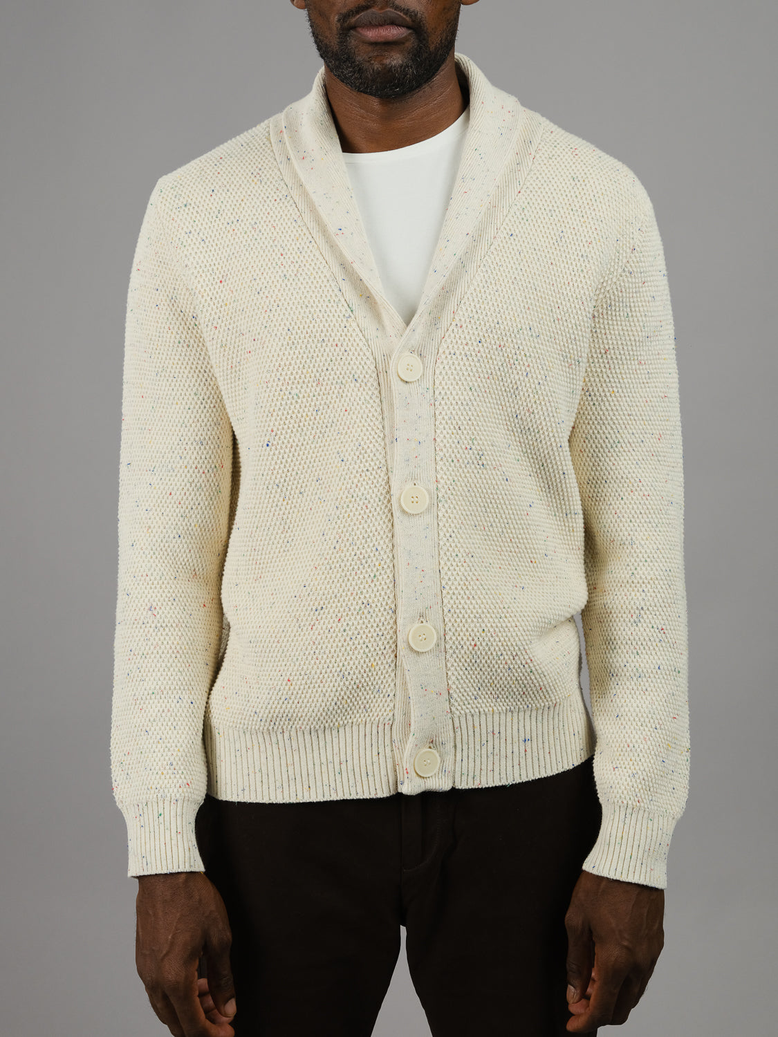 Someone sports the cream Vermont Shawl Collar Cardigan by ID Menswear, made from speckled yarn and accented with large buttons, over a white T-shirt. The plain gray background highlights the outfit while keeping the face hidden.