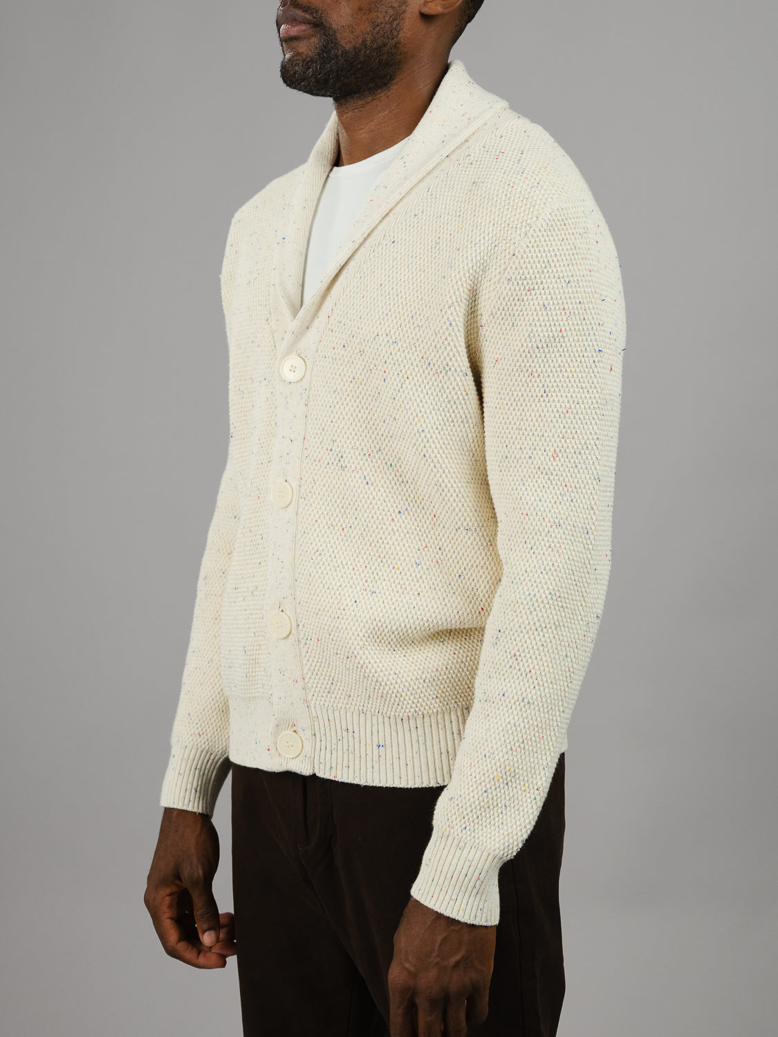 A person dons ID Menswears Vermont Shawl Collar Cardigan in cream, made from speckled yarn with large buttons, over a white shirt. Dark pants complete the look against a plain gray background.
