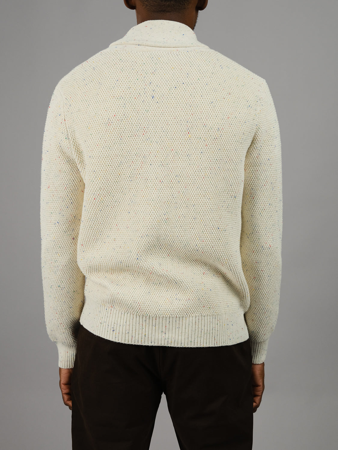 A person wearing an ID Menswear Vermont Shawl Collar Cardigan in cream, paired with dark pants, stands against a plain gray background, facing away from the camera.