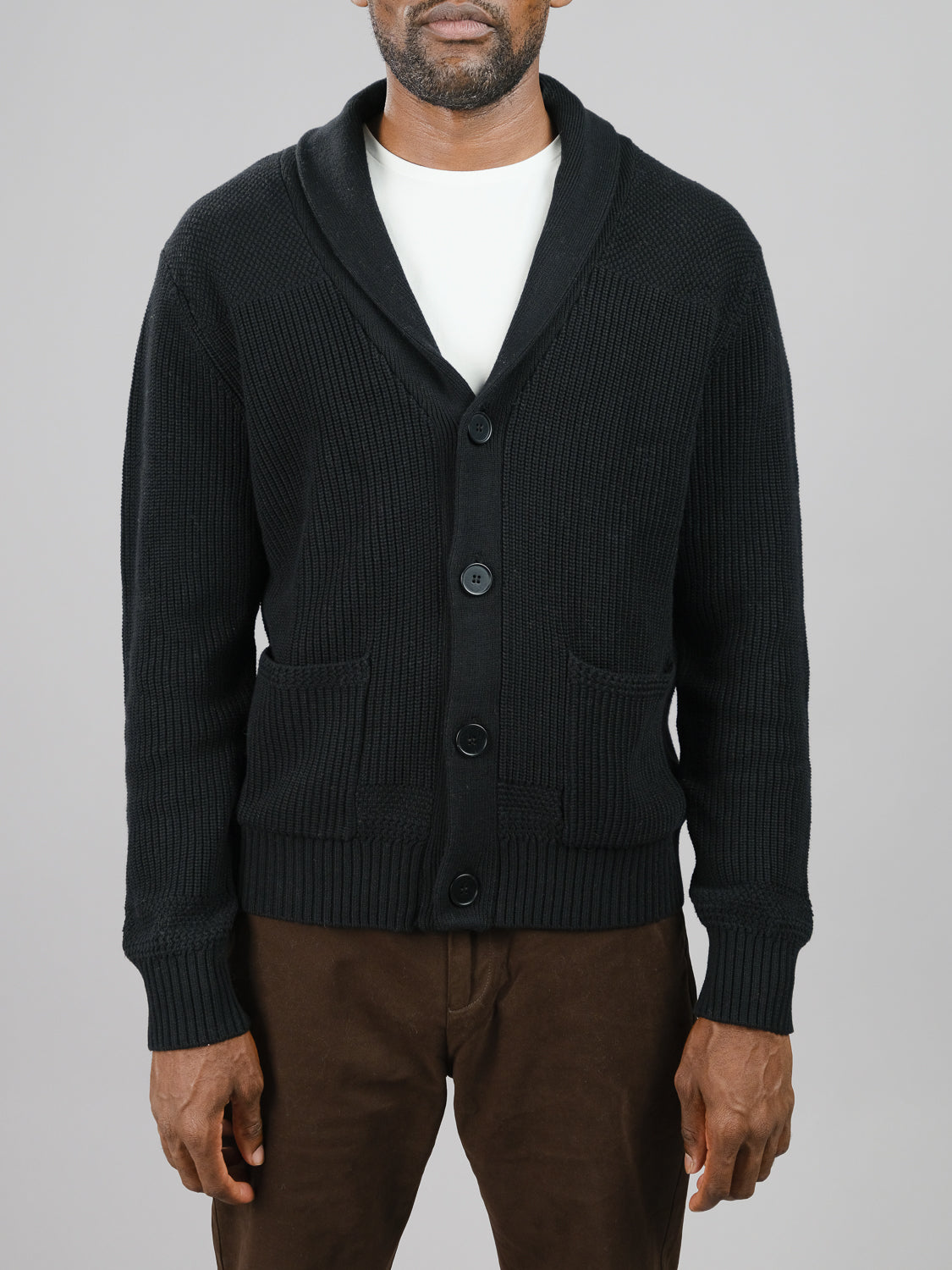 A man wears the ID Menswear Helsinki Cotton Knitted Cardigan with a classic shawl collar and buttoned front, layered over a white shirt and paired with brown pants, set against a plain gray background.