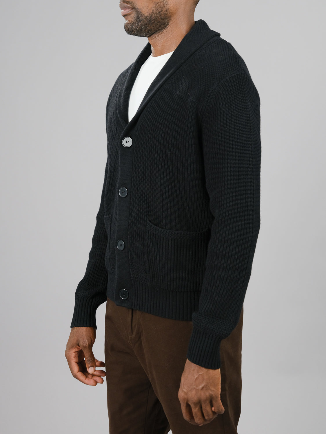 A person wears an ID Menswear black Helsinki Cotton Knitted Cardigan, featuring a classic shawl collar, ribbed texture, and pockets, over a white shirt and brown pants against a plain gray background. The persons head is not visible in the image.