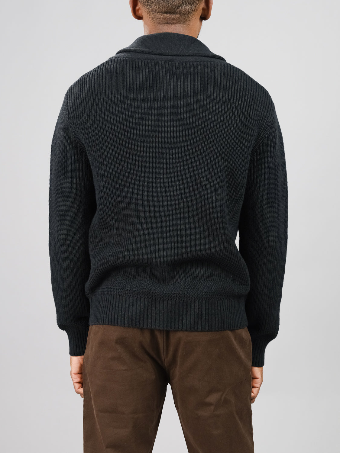 A person is seen from behind wearing an ID Menswear Helsinki Cotton Knitted Cardigan in black, featuring a classic shawl collar and long sleeves. It is paired with brown pants against a gray background, perfectly complementing their short hair.