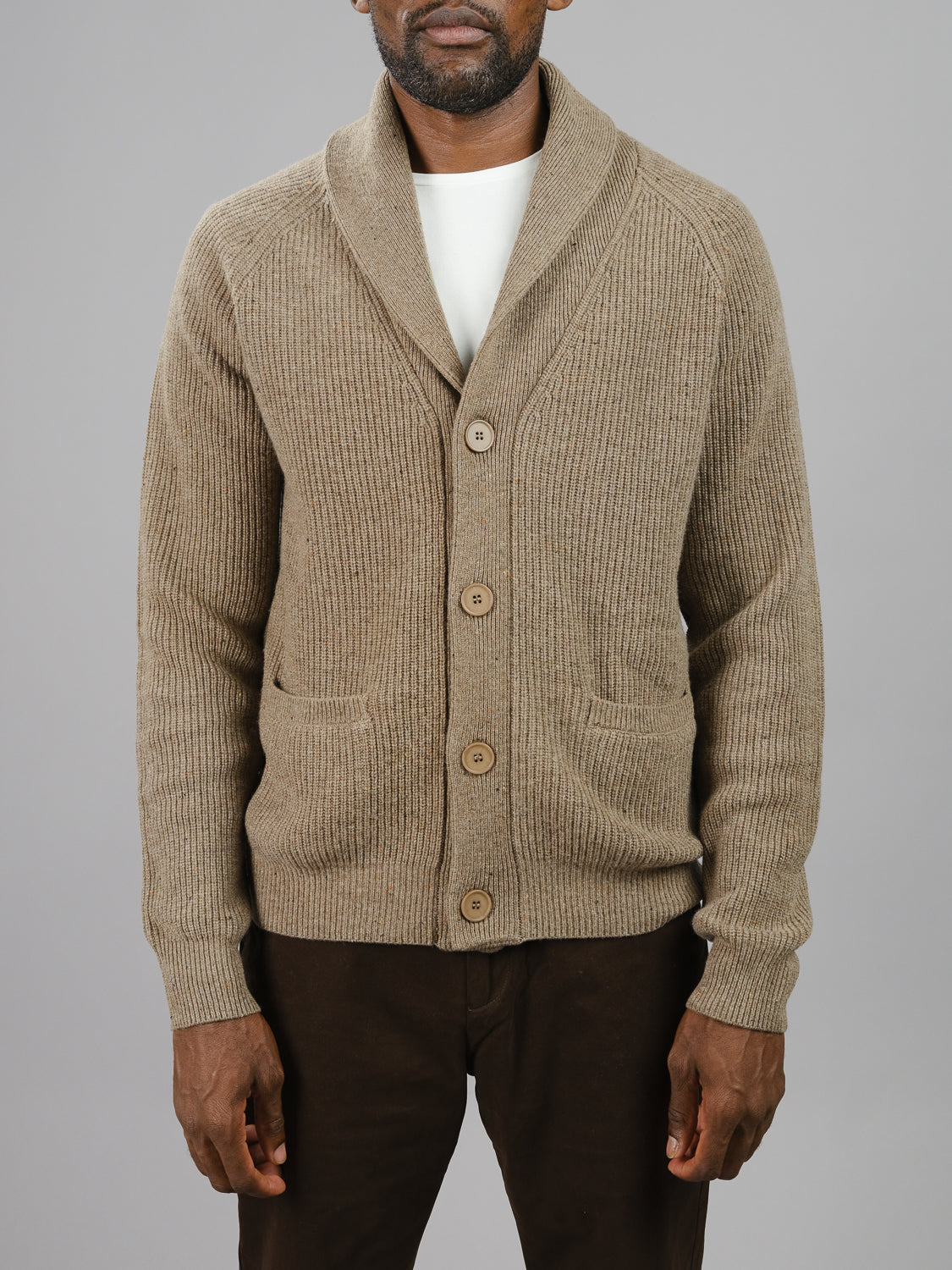 A man wears an ID Menswears Montreal Merino Wool Cardigan, with ribbed beige fabric, a shawl collar, and brown buttons over a white shirt. He pairs it with dark trousers against a plain gray backdrop.
