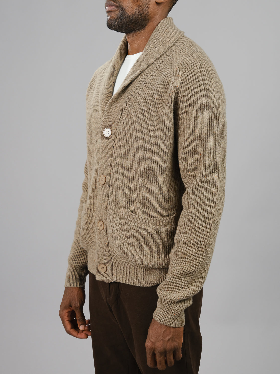 A person wears an ID Menswear Montreal Merino Wool Cardigan of speckled beige knit, featuring two pockets and a shawl collar, buttoned over a white shirt with dark brown pants against a plain gray background.
