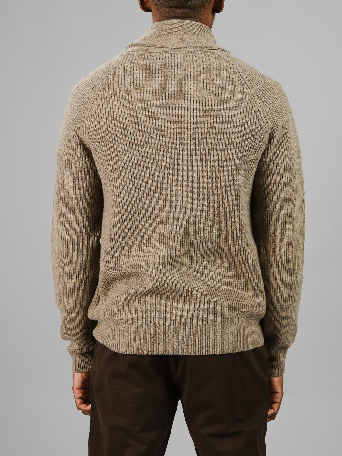 A person in a Montreal Merino Wool Cardigan by ID Menswear, featuring beige ribbed knit, and dark pants is shown from the back against a plain gray background.