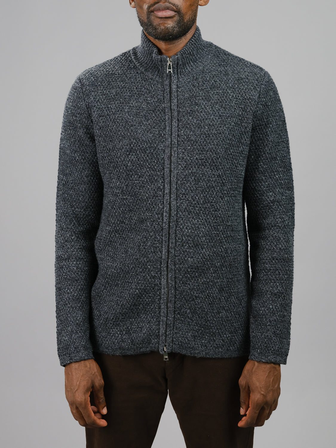 A man wears the ID Menswear Glasgow Zip Up Sweater in textured dark gray and dark brown pants, standing against a plain light gray background with his face partially visible and arms relaxed at his sides.