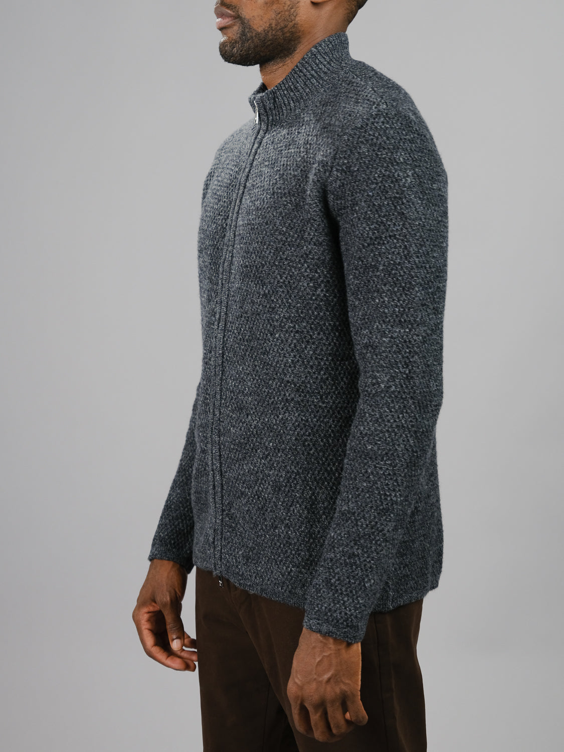 A person stands in profile against a light gray background, wearing the ID Menswear Glasgow Zip Up Sweater in textured dark gray and brown pants, with their head turned slightly to the left.