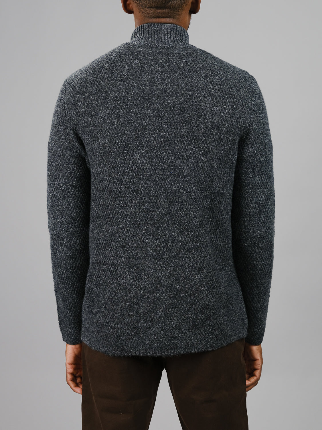 A person is shown from the back against a plain gray background wearing the ID Menswear Glasgow Zip Up Sweater in textured dark gray merino wool and brown pants.