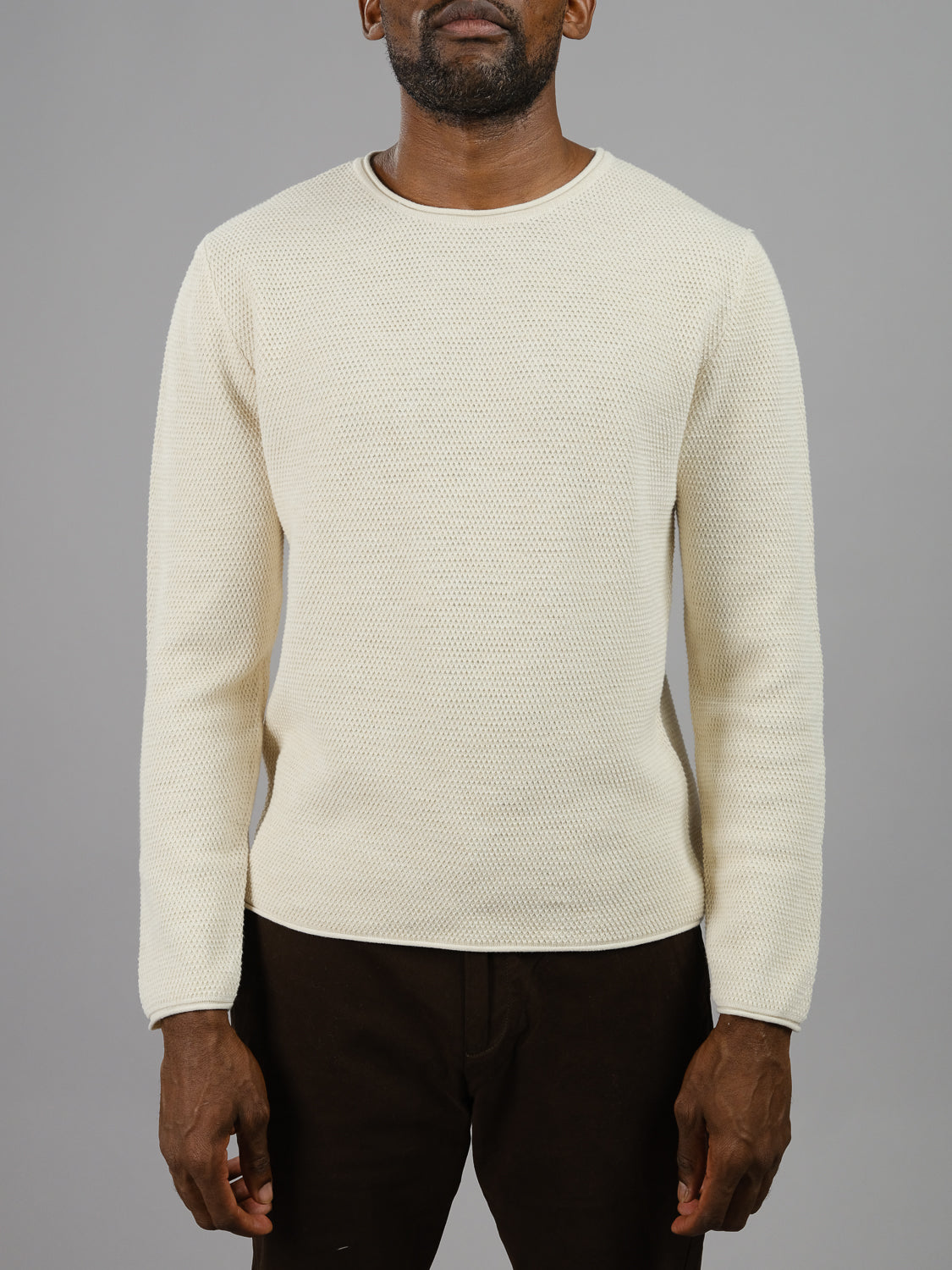 A person in a cream Malta Rolled Neck Sweater Tee from ID Menswear stands against a gray background. The long-sleeve, textured tee, made of soft cotton, offers a comfortable fit with a round neckline and subtle woven pattern. Their arms rest relaxed by their sides.