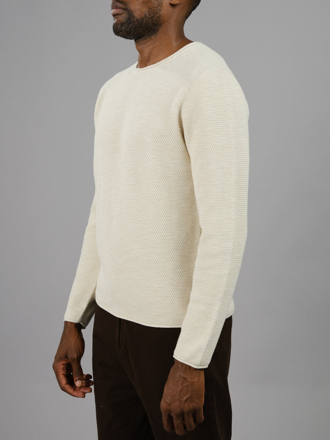 The image features a person in an off-white Malta Rolled Neck Sweater Tee by ID Menswear, coupled with dark pants, against a gray backdrop. The side profile highlights the cozy fit and textured detail of the sweater.