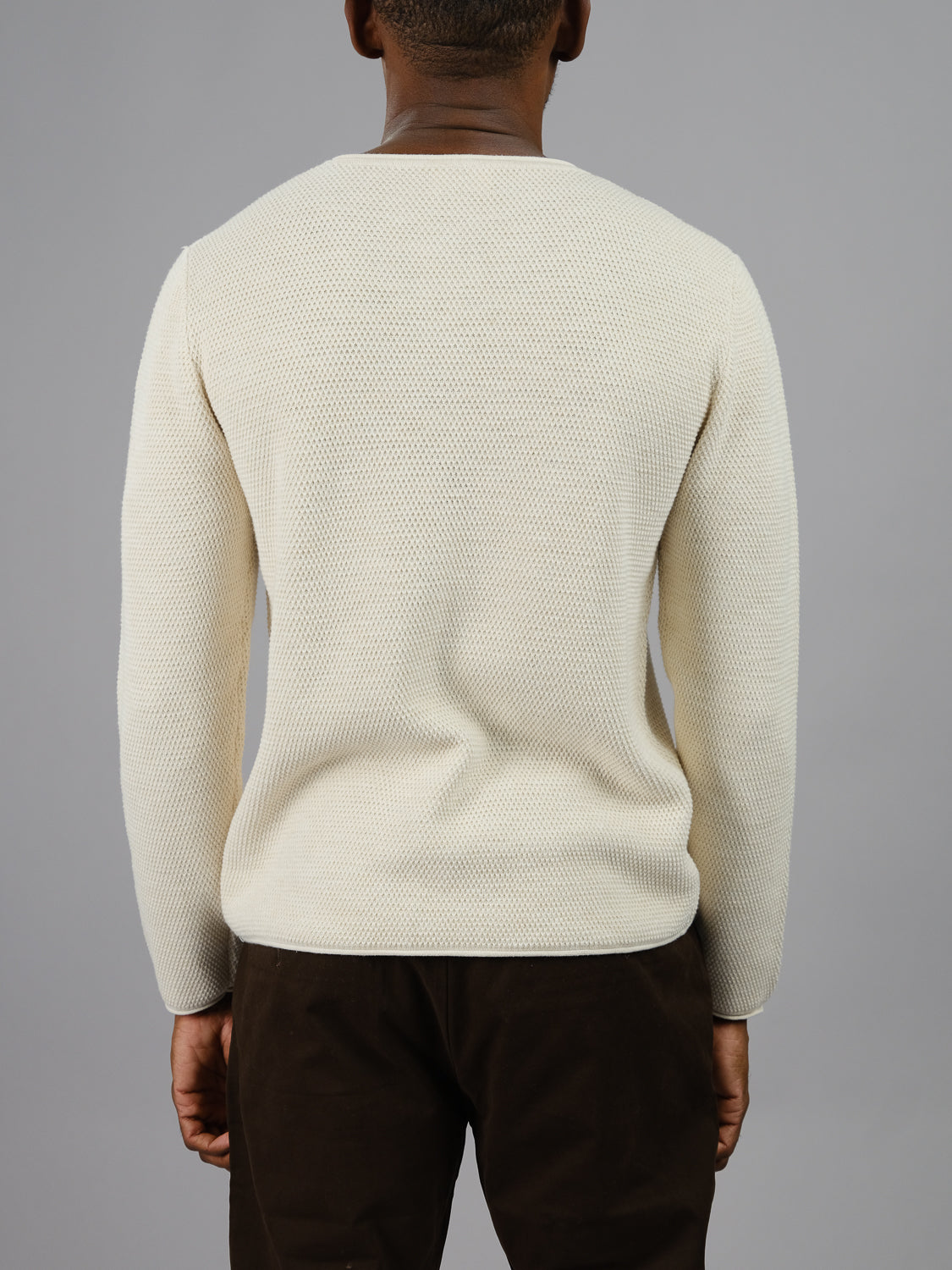 A person stands with their back to the camera in an ID Menswear Malta Rolled Neck Sweater Tee, featuring a textured cream design and dark pants, against a plain gray background.