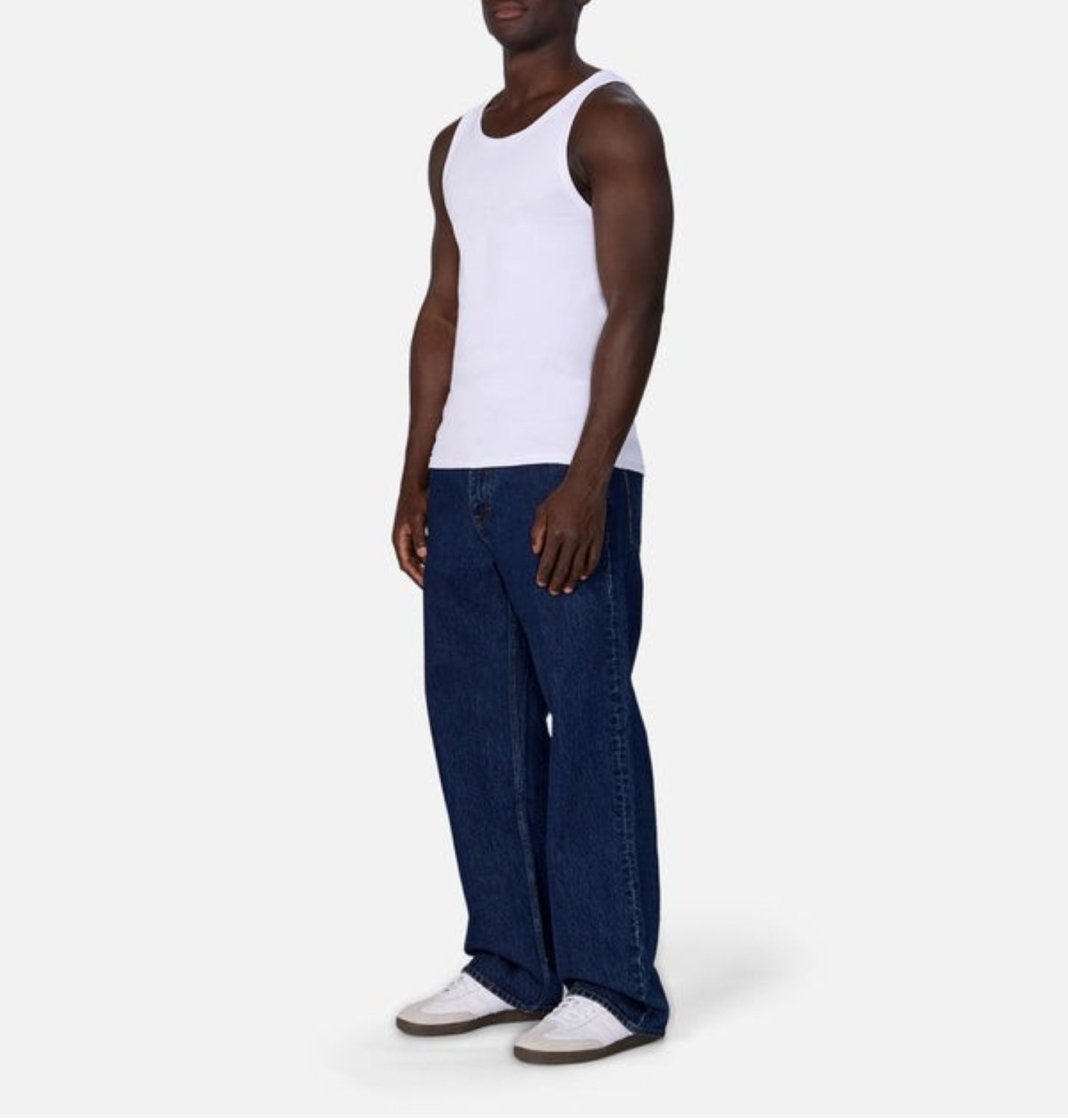 A man stands against a plain background wearing the Washed Brando Ribbed Tank Top by Industrie, paired with loose blue jeans and white sneakers—an effortless summer staple.