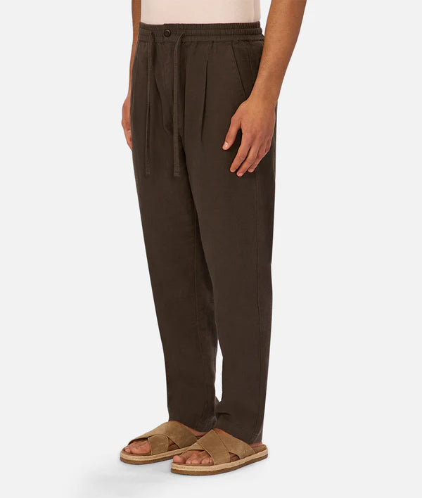 A person wears The Mallorca Pant by Industrie, featuring loose-fitting cotton in dark brown with pleats and a breathable design, paired with tan sandals. The image shows only legs and feet against a white background.