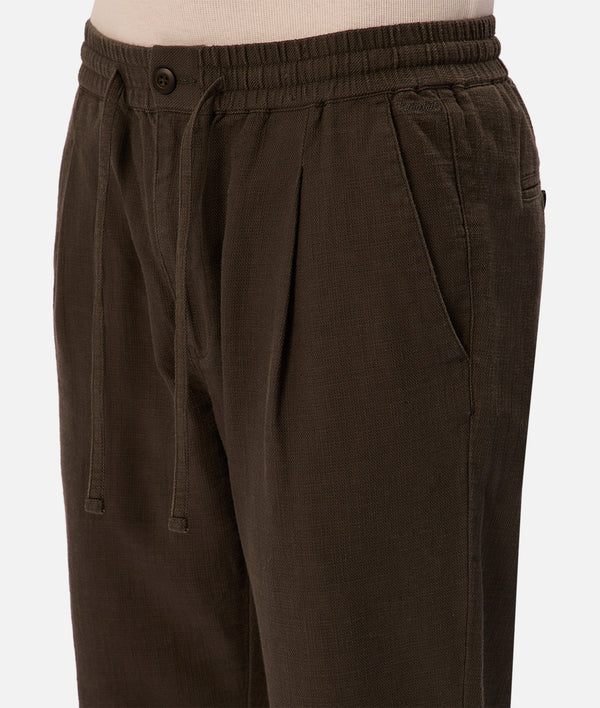 Close-up of a person wearing The Mallorca Pant by Industrie—breathable cotton pants with an elastic waistband, button closure, side pockets, and subtle pleats in dark brown.