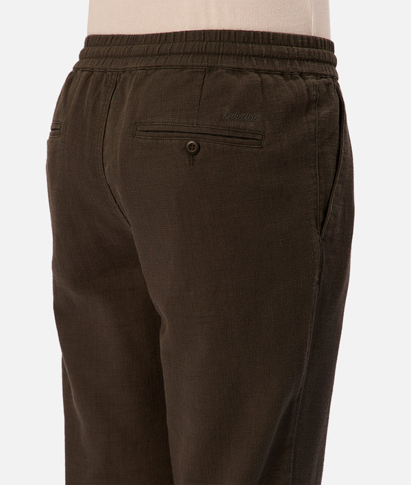 A close-up of a person wearing Industrie’s The Mallorca Pant in dark brown, featuring breathable cotton, an elastic waistband, side pockets, and a buttoned back pocket. The upper part of a beige shirt is also visible.
