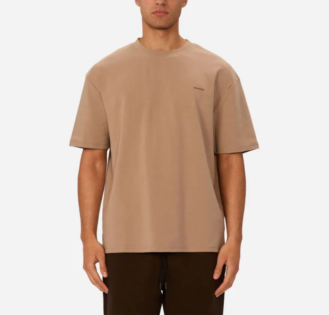 A person wearing Industrie’s The Newport T-Shirt, a loose-fitting beige 90s-inspired oversized tee with a small left chest logo, paired with dark pants and standing against a plain white background.