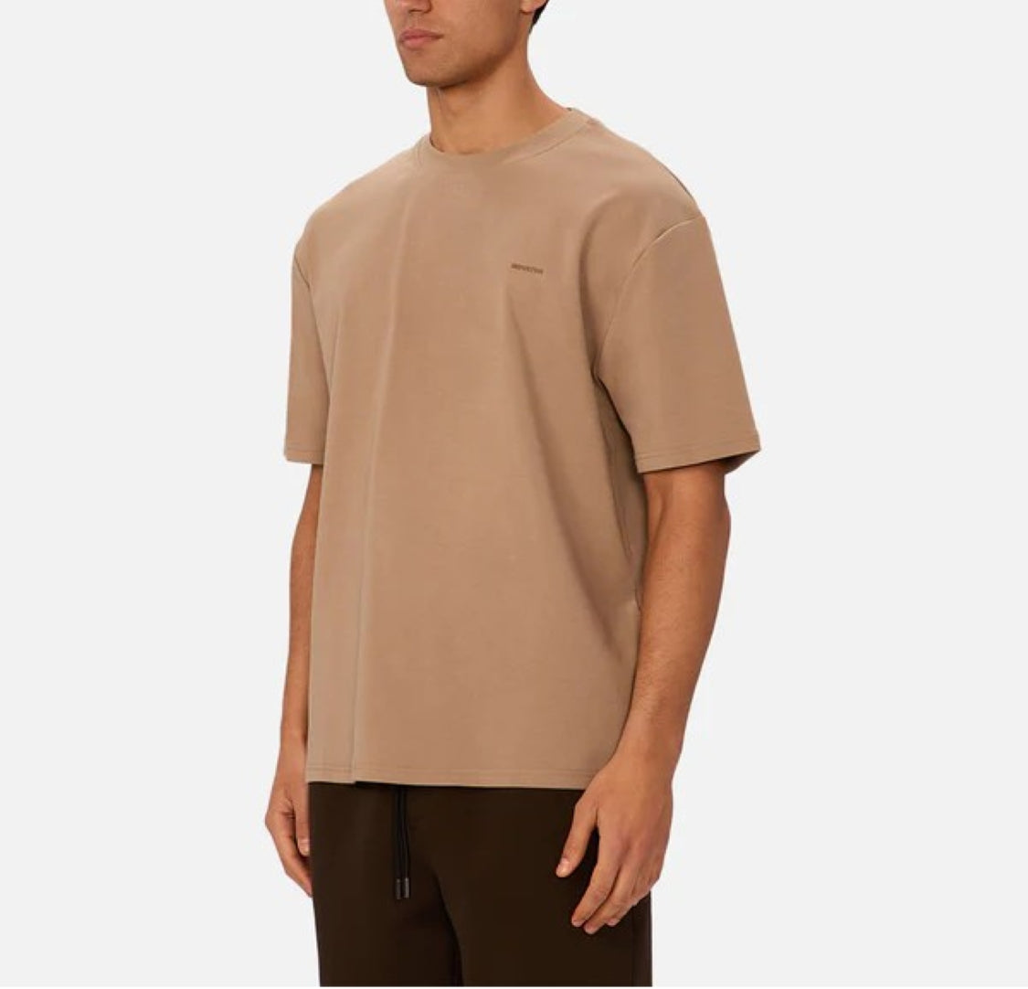 A person wearing Industrie’s The Newport T-Shirt, a loose-fitting, 90s-inspired beige oversized tee, pairs it with dark pants and stands against a plain light background, facing slightly to the side.