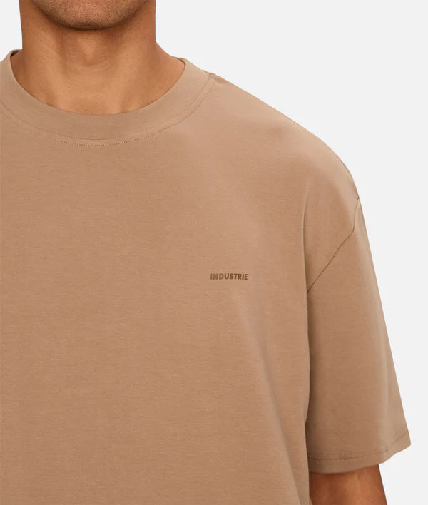 A person wears The Newport T-Shirt by Industrie—a loose, heavyweight beige crew neck with a small INDUSTRIE logo on the left chest. The 90s-inspired oversized tee is shown from shoulders to waist, with the person's head partially visible.