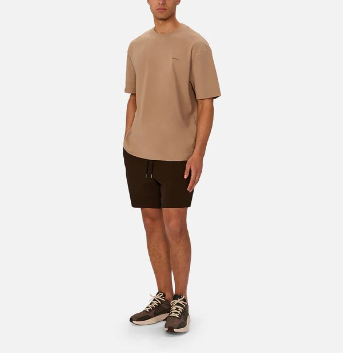 A person stands against a plain background wearing Industrie’s The Newport T-Shirt, a 90s inspired, oversized beige top paired with dark brown shorts and beige and brown sneakers.