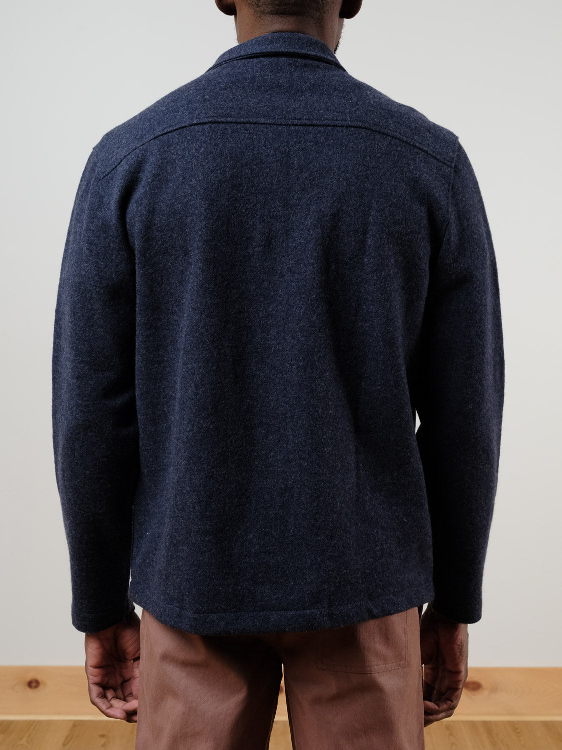 A person stands facing away from the camera, wearing the Marine Layer Mendocino Wool Overshirt in dark blue and light brown pants. Their hands are relaxed at their sides against a plain, light-colored background.