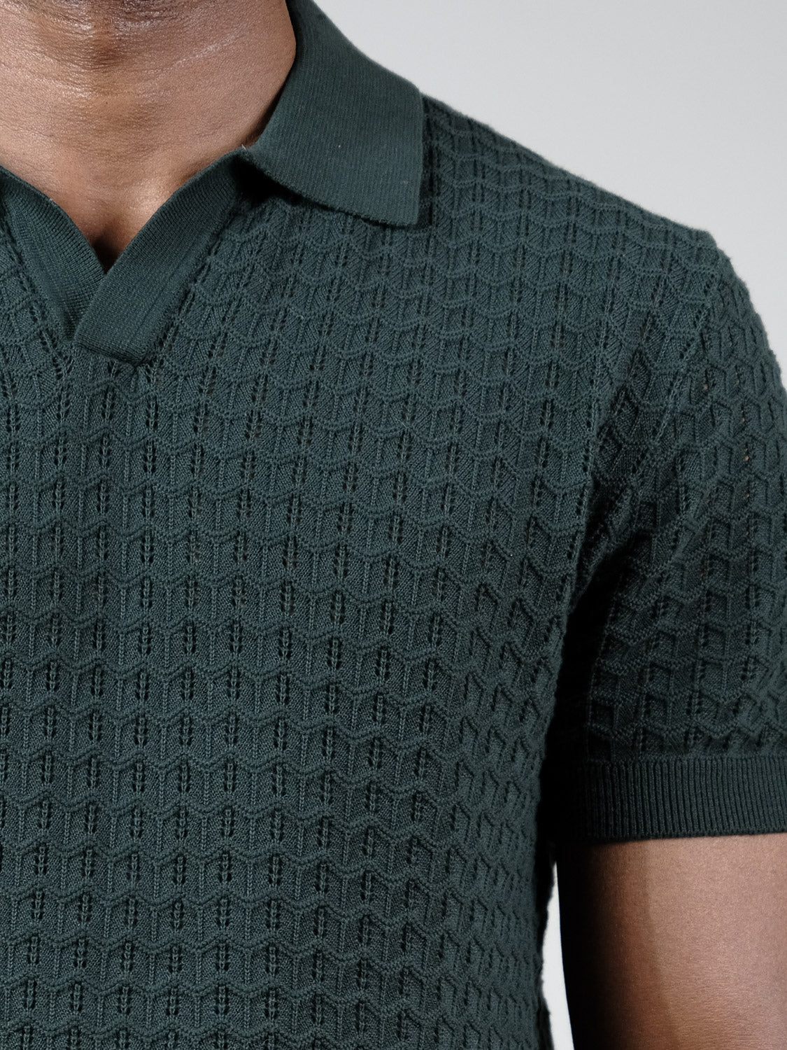 A close-up of a person wearing the Mojave Knitted Cotton Polo by ID Menswear—a dark green, short-sleeve collared shirt with a geometric pattern. Only the lower face, neck, and upper torso are visible; ideal for casual wear.