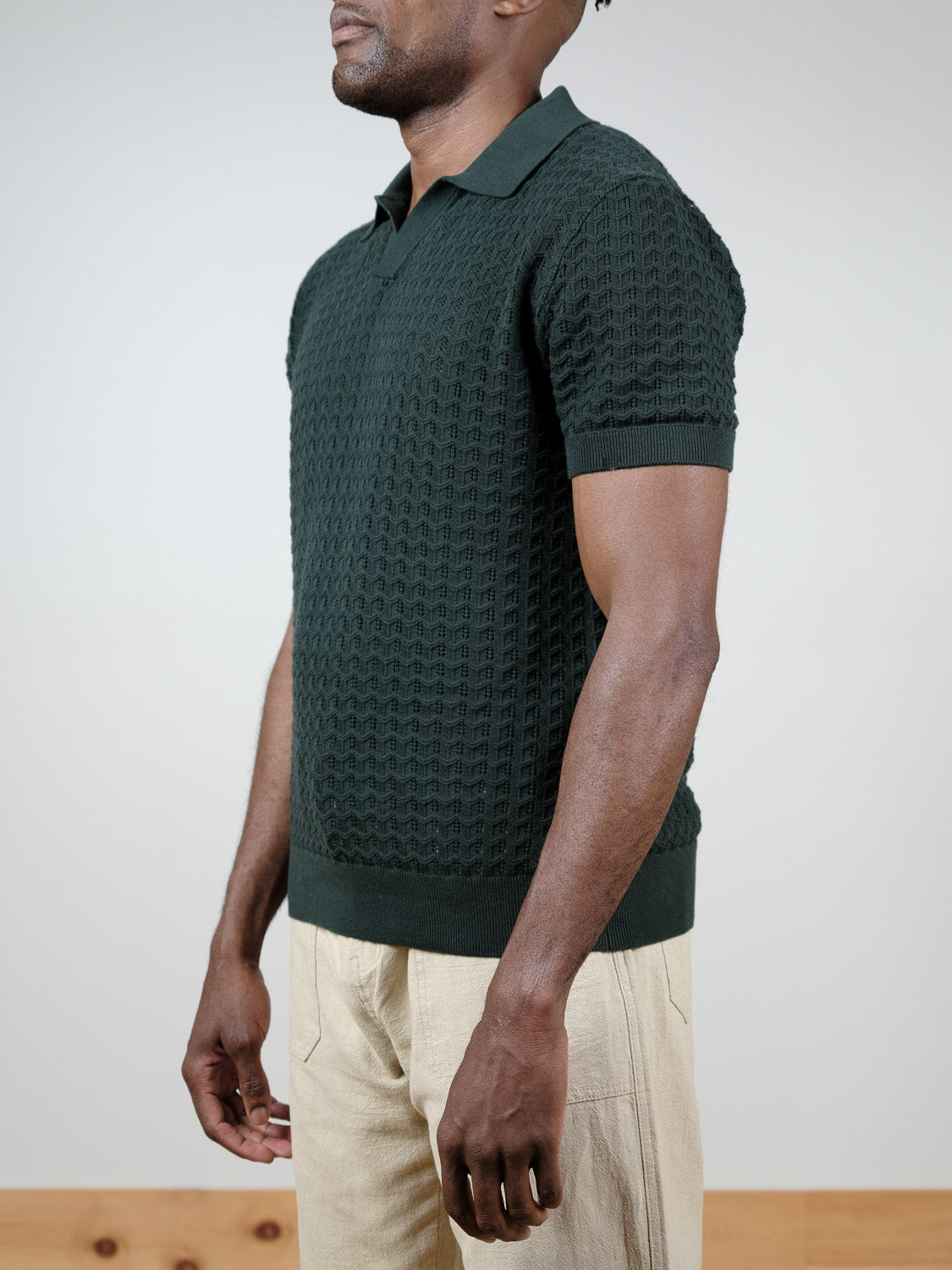 A man poses against a plain background, wearing the Mojave Knitted Cotton Polo by ID Menswear in dark green with light beige pants. His head is turned slightly to the side, embodying effortless casual style.