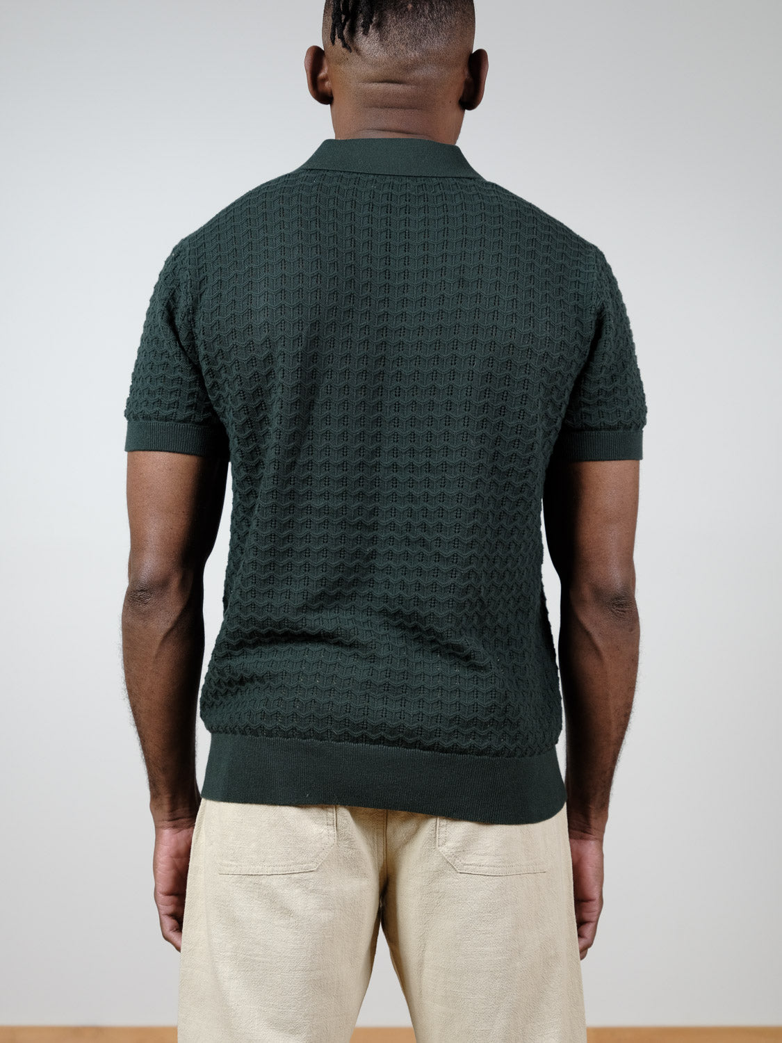 A person faces away from the camera, wearing the textured dark green Mojave Knitted Cotton Polo by ID Menswear and light beige pants, set against a plain light background.