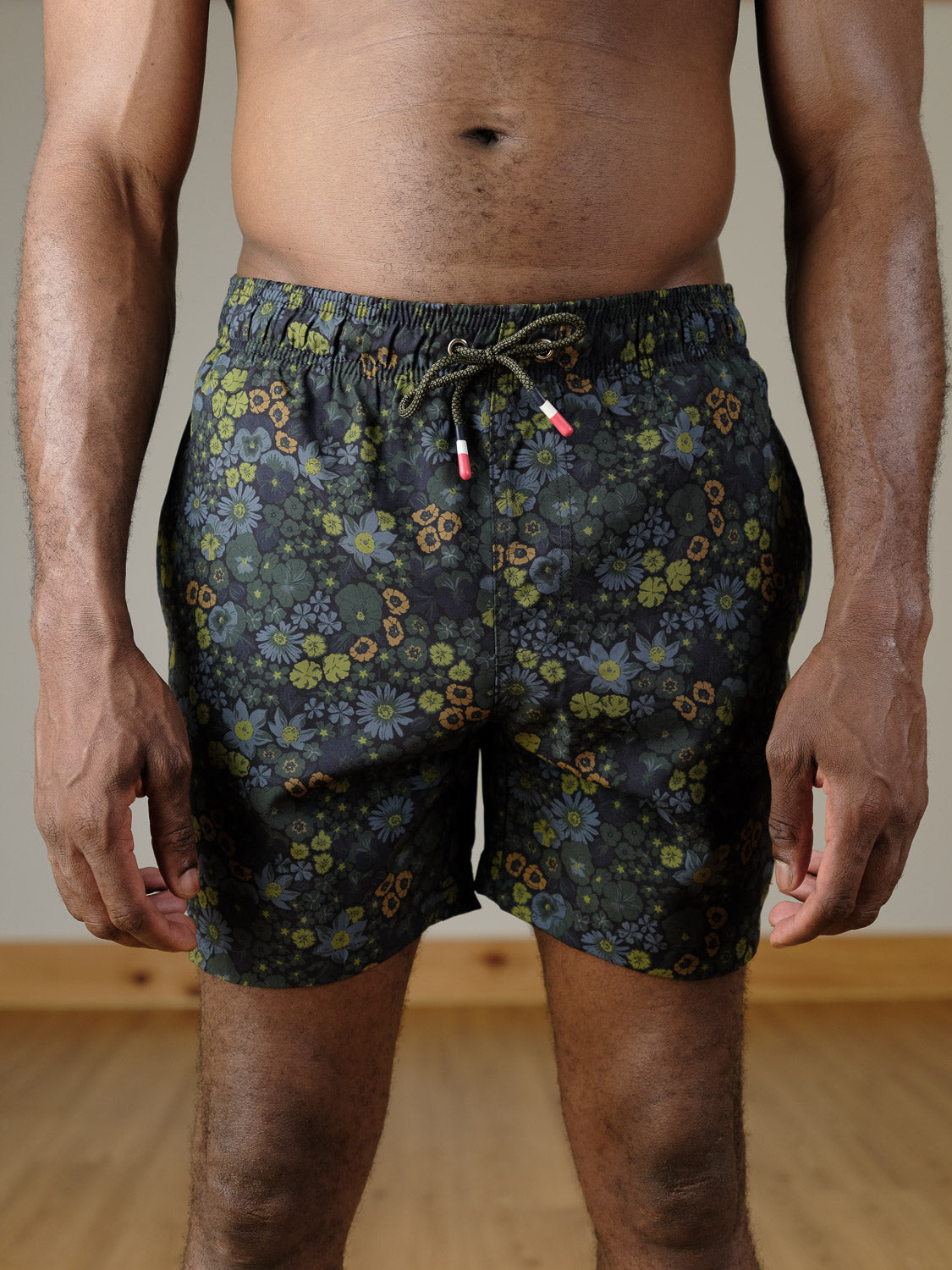 A person stands indoors sporting ID Menswear’s Wild Flower Fast Dry 4-Way Stretch Swim Trunk, featuring a dark floral pattern and drawstring, shown from waist to mid-thigh against a wood floor and neutral wall.