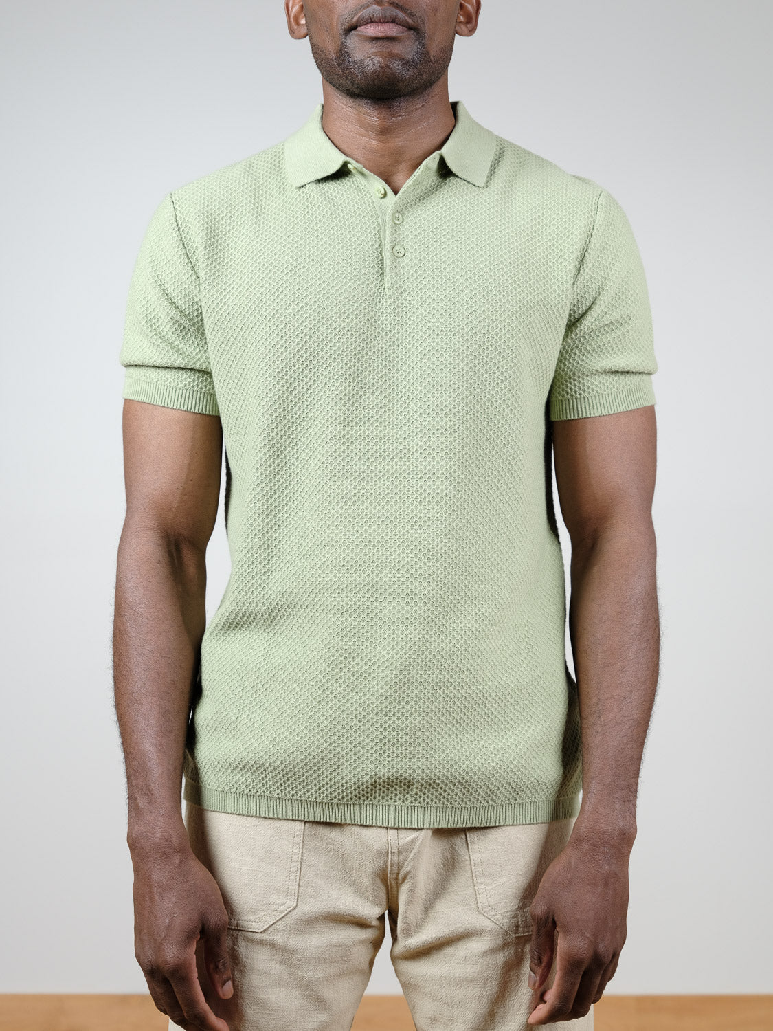 A man faces forward wearing the ID Menswear Tucson Knitted Cotton Polo in light green with beige pants against a plain light gray background. His face is partially out of frame.