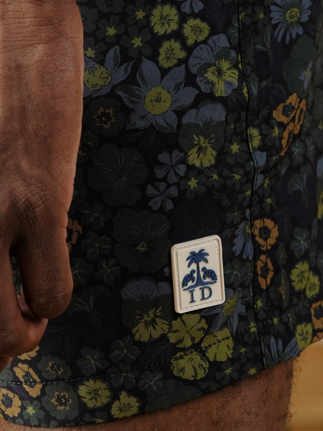 A close-up of a hand resting beside ID Menswear's Wild Flower Fast Dry 4-Way Stretch Swim Trunk, featuring a dark floral pattern and a rectangular patch with palm trees and "ID.