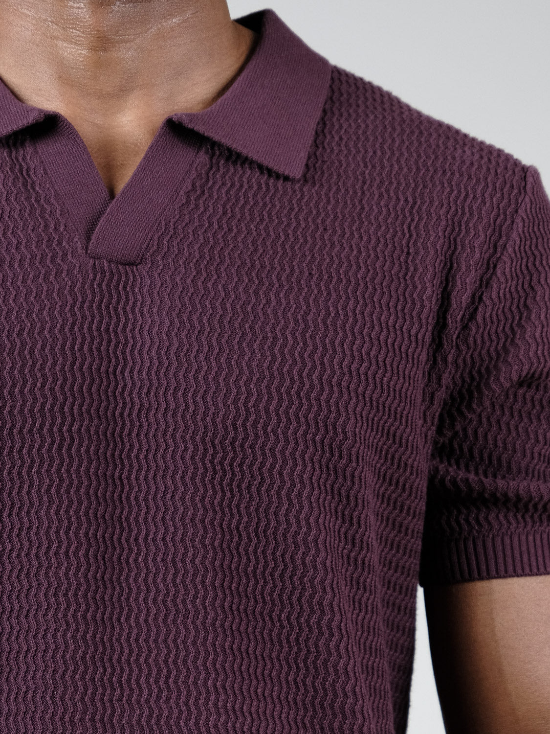 A close-up shows a person wearing the Mecca Knitted Cotton Polo by ID Menswear, featuring a structured collar, open neckline, textured short sleeves, and dark purple cotton that highlights the upper chest and shoulders.