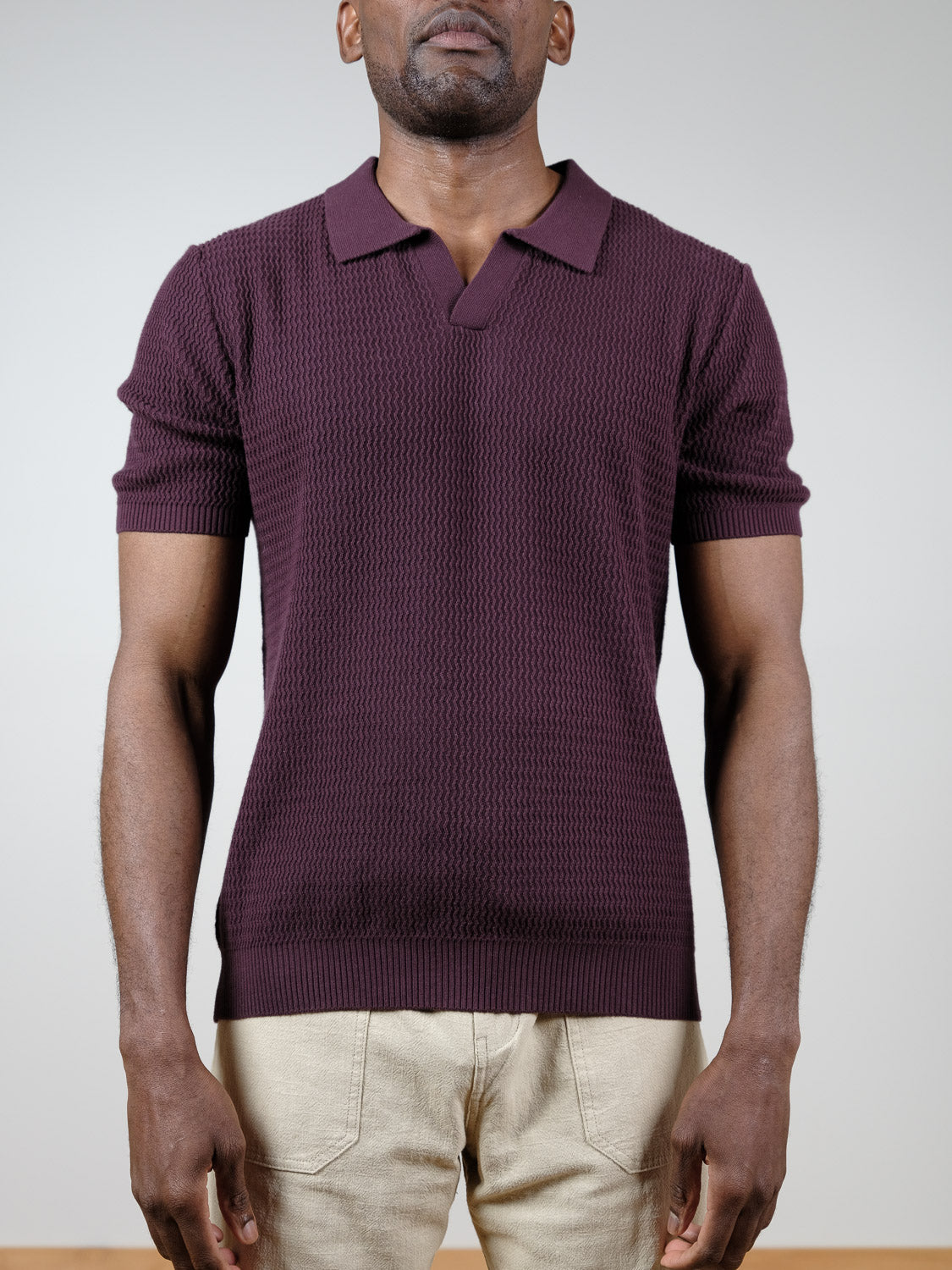 A man wears the Mecca Knitted Cotton Polo by ID Menswear in burgundy with light beige pants, standing against a plain light background. His face is partially out of the frame.