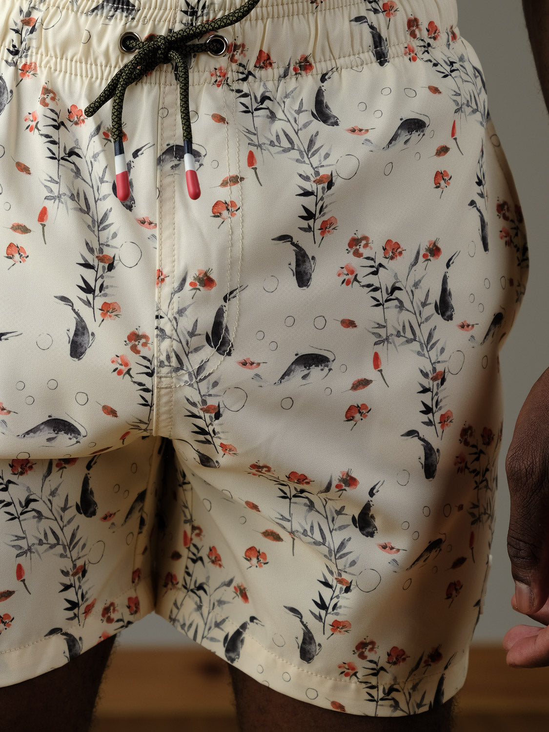 Close-up of a person wearing the Koi Fast Dry 4-Way Stretch Swim Trunk by ID Menswear, with a playful print of whales, fish, bubbles, red flowers, and green leaves. Green drawstring has red and white tips; part of the hand is visible.