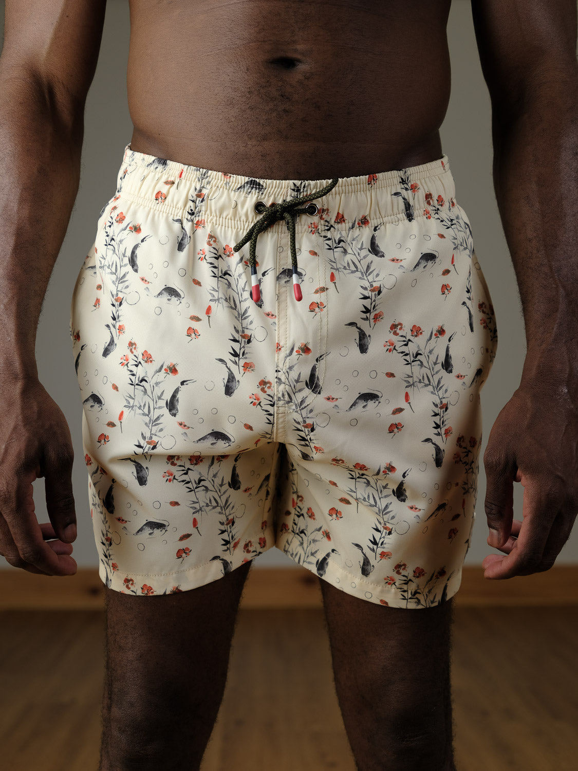 A person stands indoors on a wooden floor wearing ID Menswear's Koi Fast Dry 4-Way Stretch Swim Trunk, featuring a light-colored fabric with a floral and fish pattern. The image is cropped at the chest and above the knees.