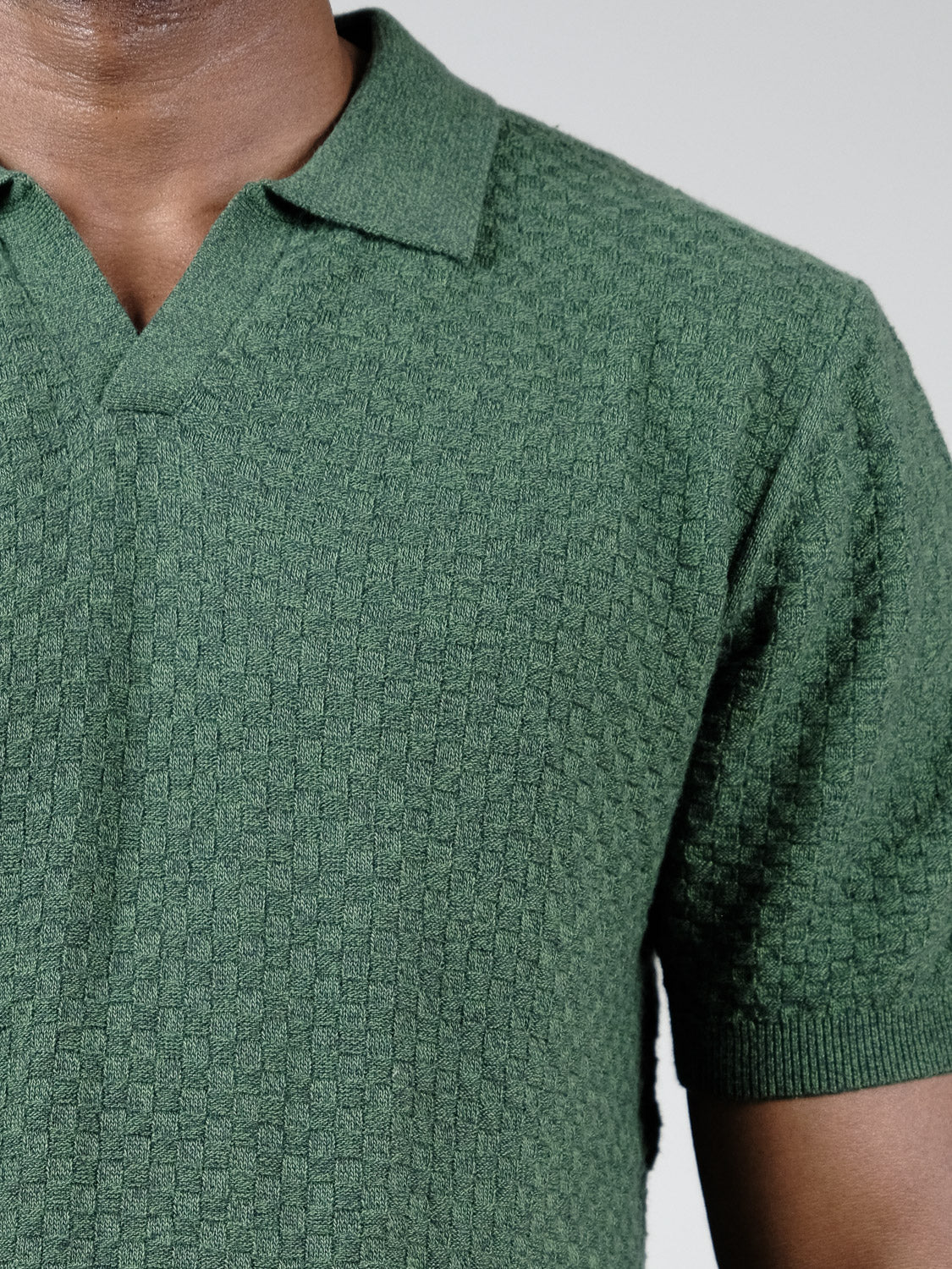 A close-up of a person wearing ID Menswear's Cairo Knitted Cotton Polo, featuring a textured green short-sleeve design with collar and v-neck, highlights the breathable knit against a plain light gray background.