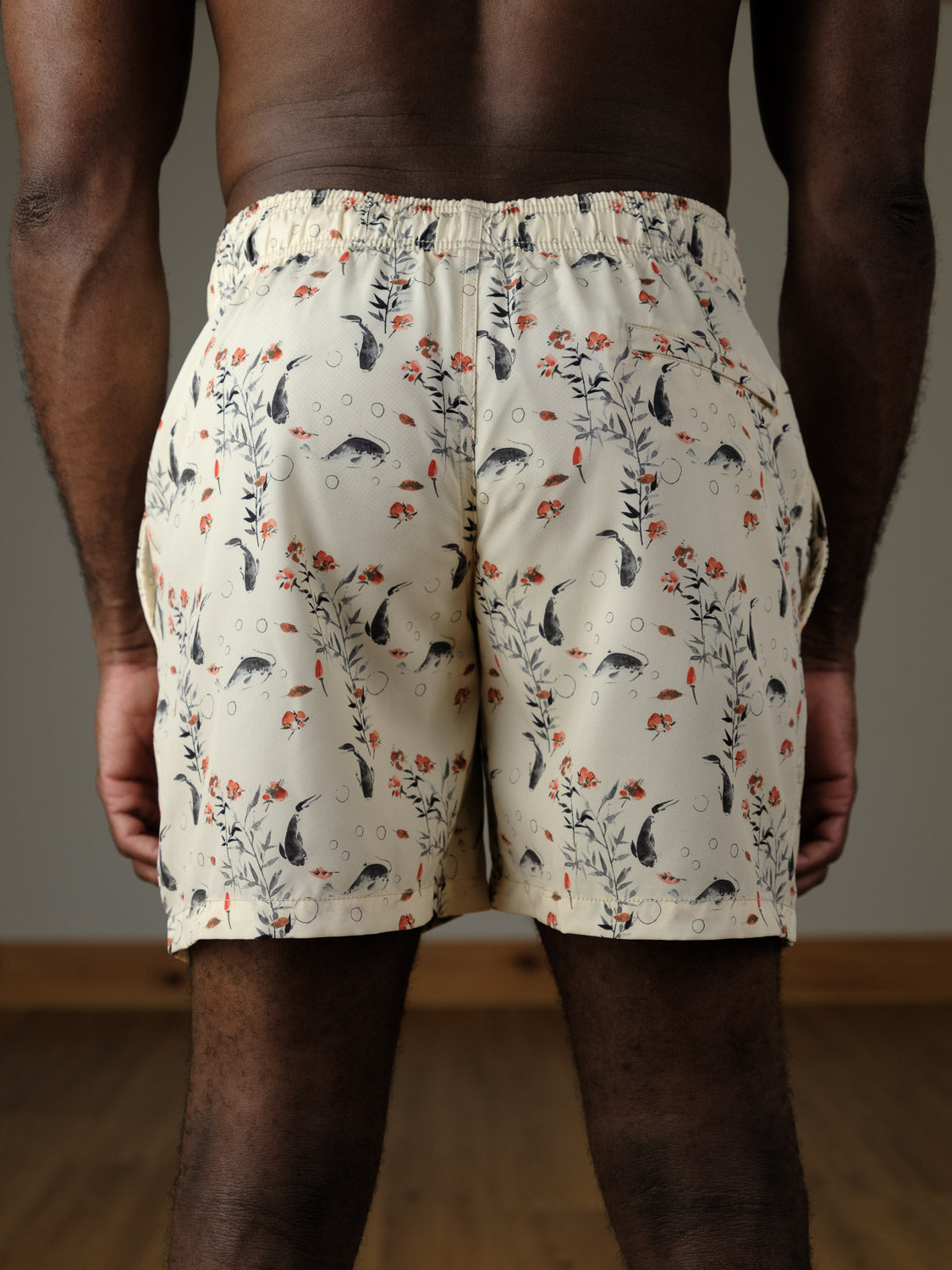 A person faces away, wearing ID Menswear's Koi Fast Dry 4-Way Stretch Swim Trunk, featuring a floral and fish print, quick-dry fabric, and elastic waistband. The softly lit scene includes a wood floor and plain wall.