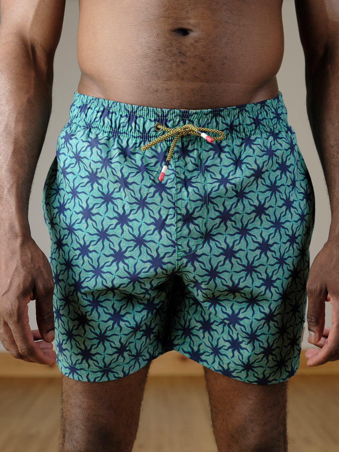 A person stands indoors wearing ID Menswear's Arica Fast Dry 4-Way Stretch Swim Trunk in blue and green with a starfish pattern. The focus is on the shorts and torso, hands resting at their sides.