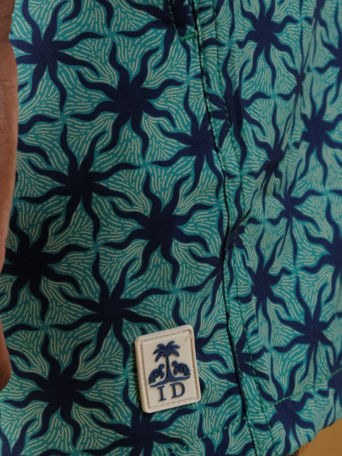 A close-up of the Arica Fast Dry 4-Way Stretch Swim Trunk by ID Menswear shows blue and turquoise starburst fabric with a white patch featuring palm trees, coconuts, and "ID"—crafted from quick-dry material for water comfort.