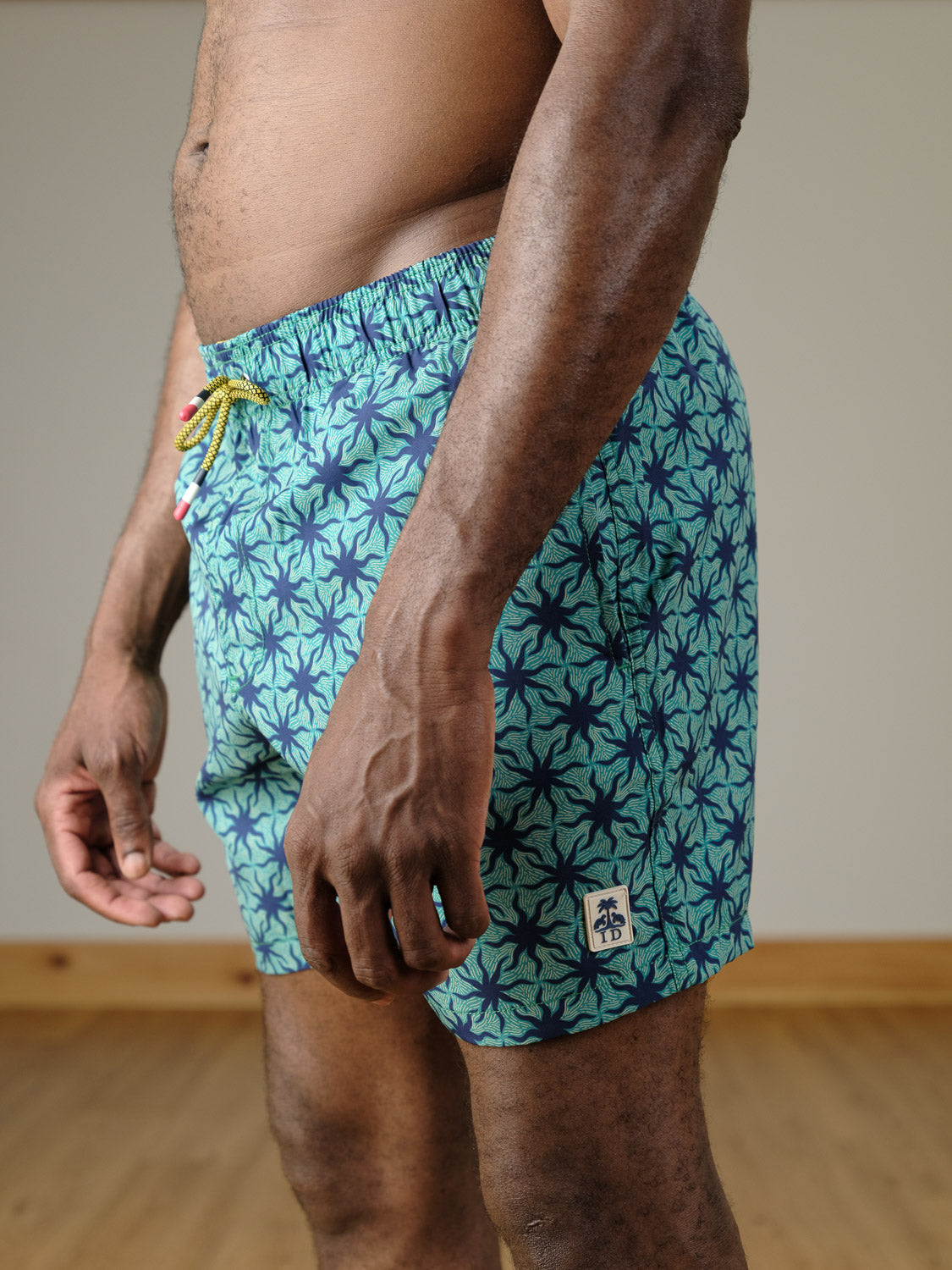 A person is seen indoors on a wooden floor wearing the Arica Fast Dry 4-Way Stretch Swim Trunk from ID Menswear. Only their lower torso, hips, and upper legs are visible.