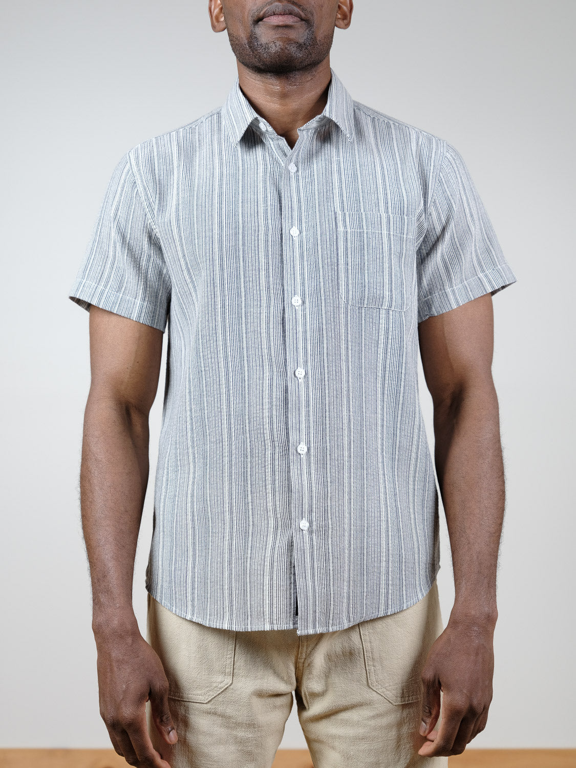 A man wears the Strider Short Sleeve Shirt by ID Menswear—relaxed fit, light gray and white stripes—with beige pants, standing against a plain light background. His face is partially out of frame.