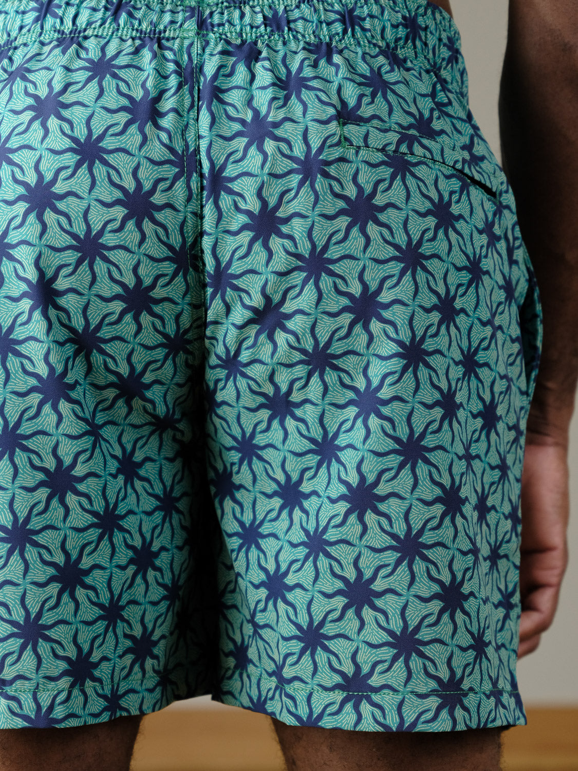 Close-up of a person wearing ID Menswear's Arica Fast Dry 4-Way Stretch Swim Trunk in teal, featuring a dark blue starfish pattern, elastic waistband, side pocket, and quick-dry fabric. Only the lower back and part of the arm are visible.