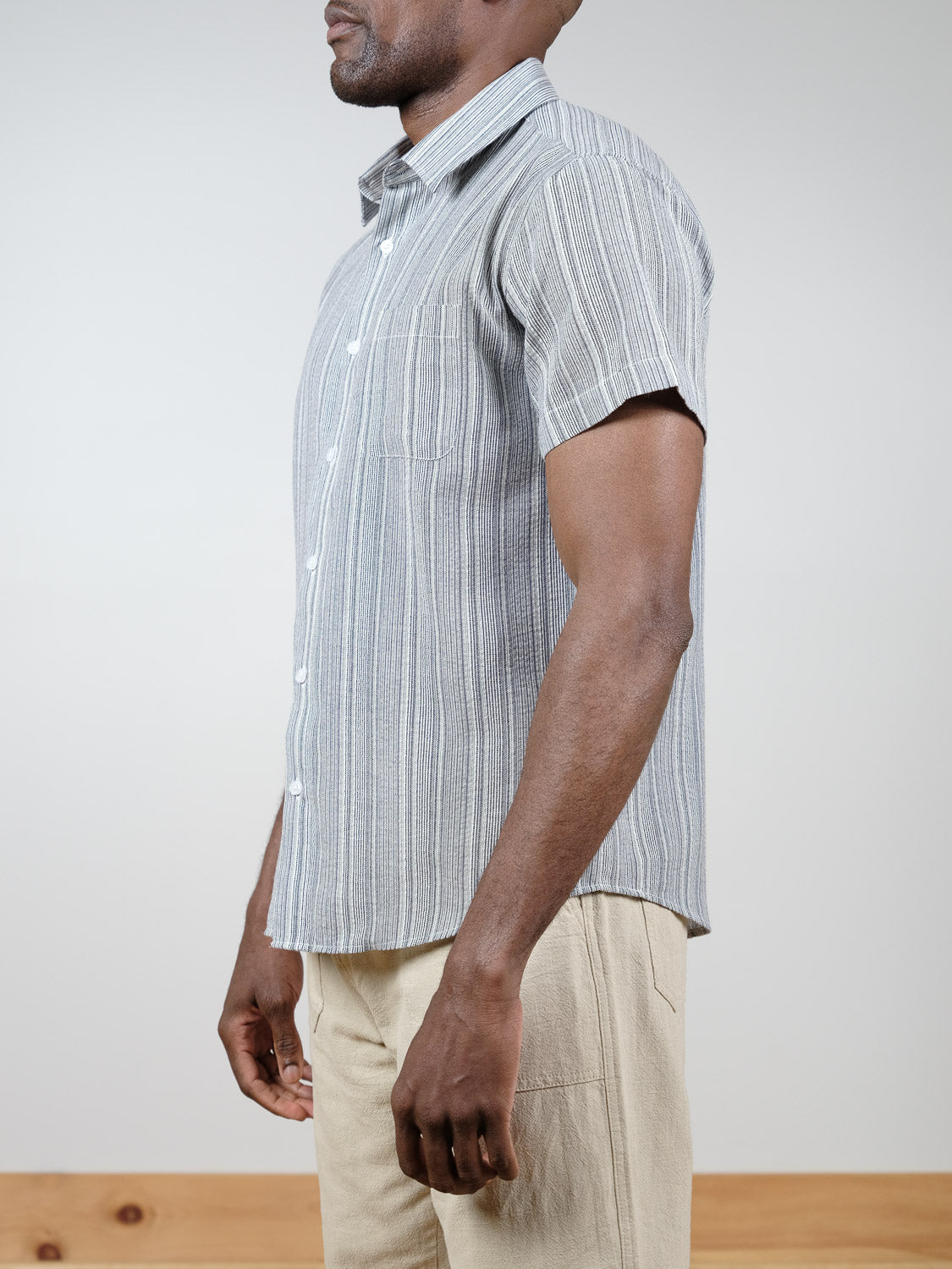 A man in profile wears the Strider Short Sleeve Shirt by ID Menswear, a relaxed-fit gray striped button-down, paired with light beige pants against a plain background.