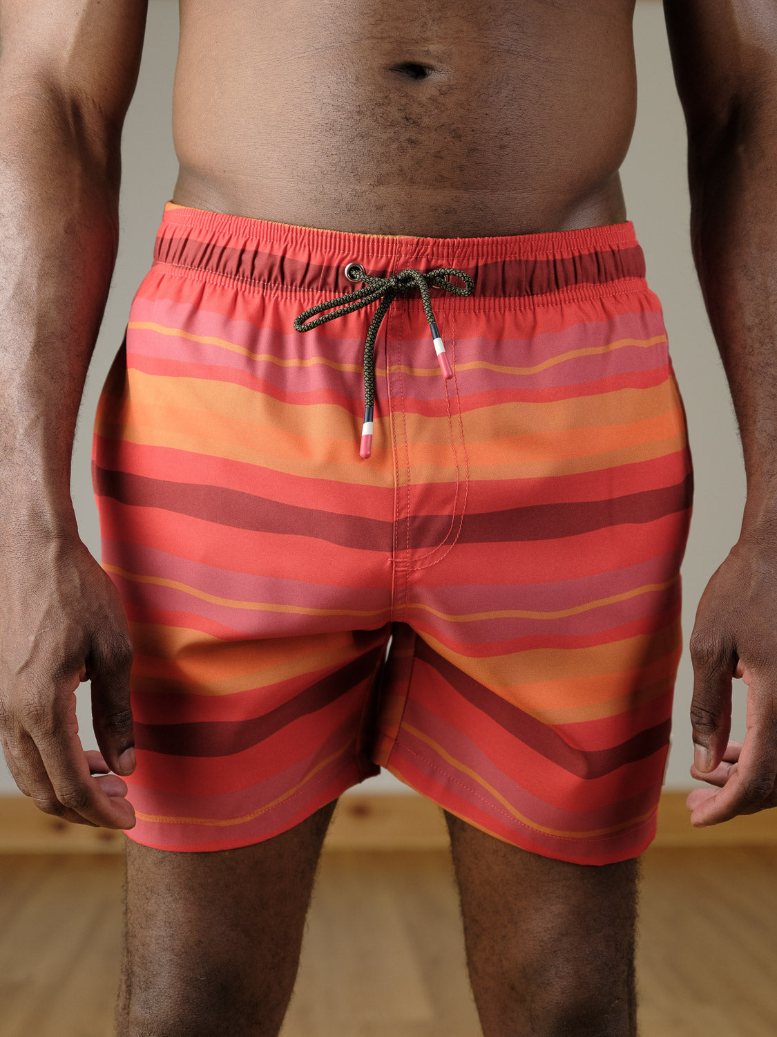 A person stands indoors on a wooden floor, shown waist to mid-thigh, wearing the ID Menswear Kochi Fast Dry 4-Way Stretch Swim Trunk in red and orange stripes with a black drawstring.