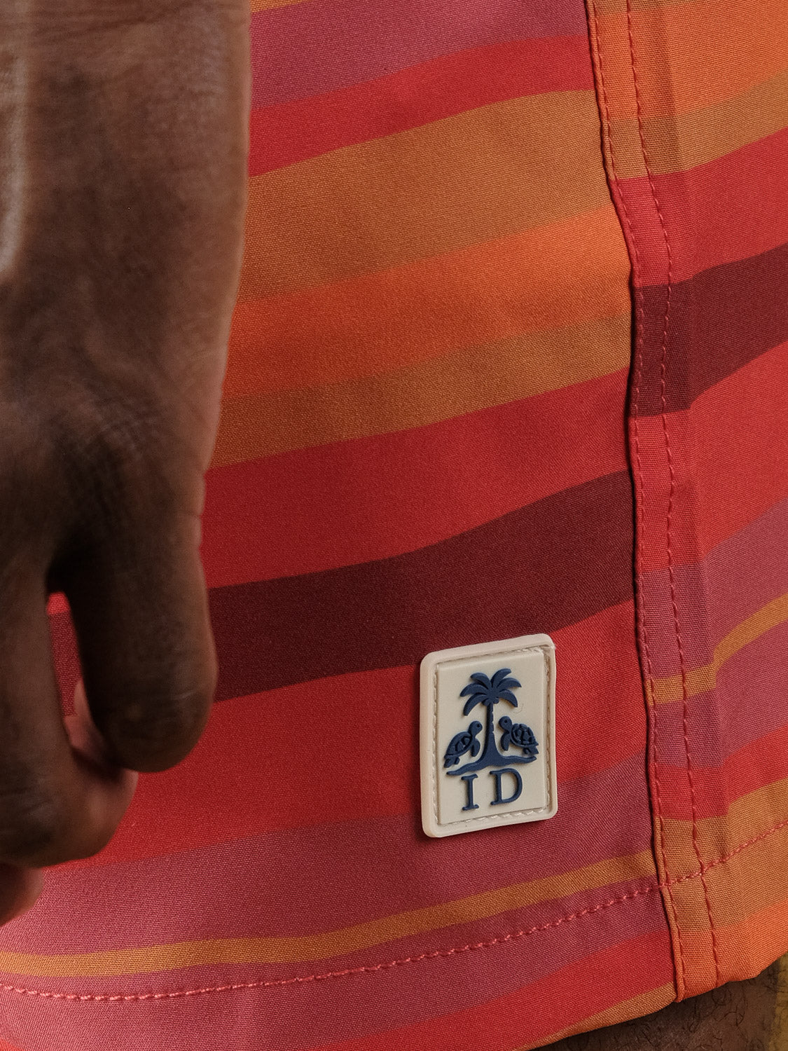 A close-up of a hand beside red and orange striped Kochi Fast Dry 4-Way Stretch Swim Trunk by ID Menswear, featuring a small patch with two birds, a palm tree, and the letters "ID.