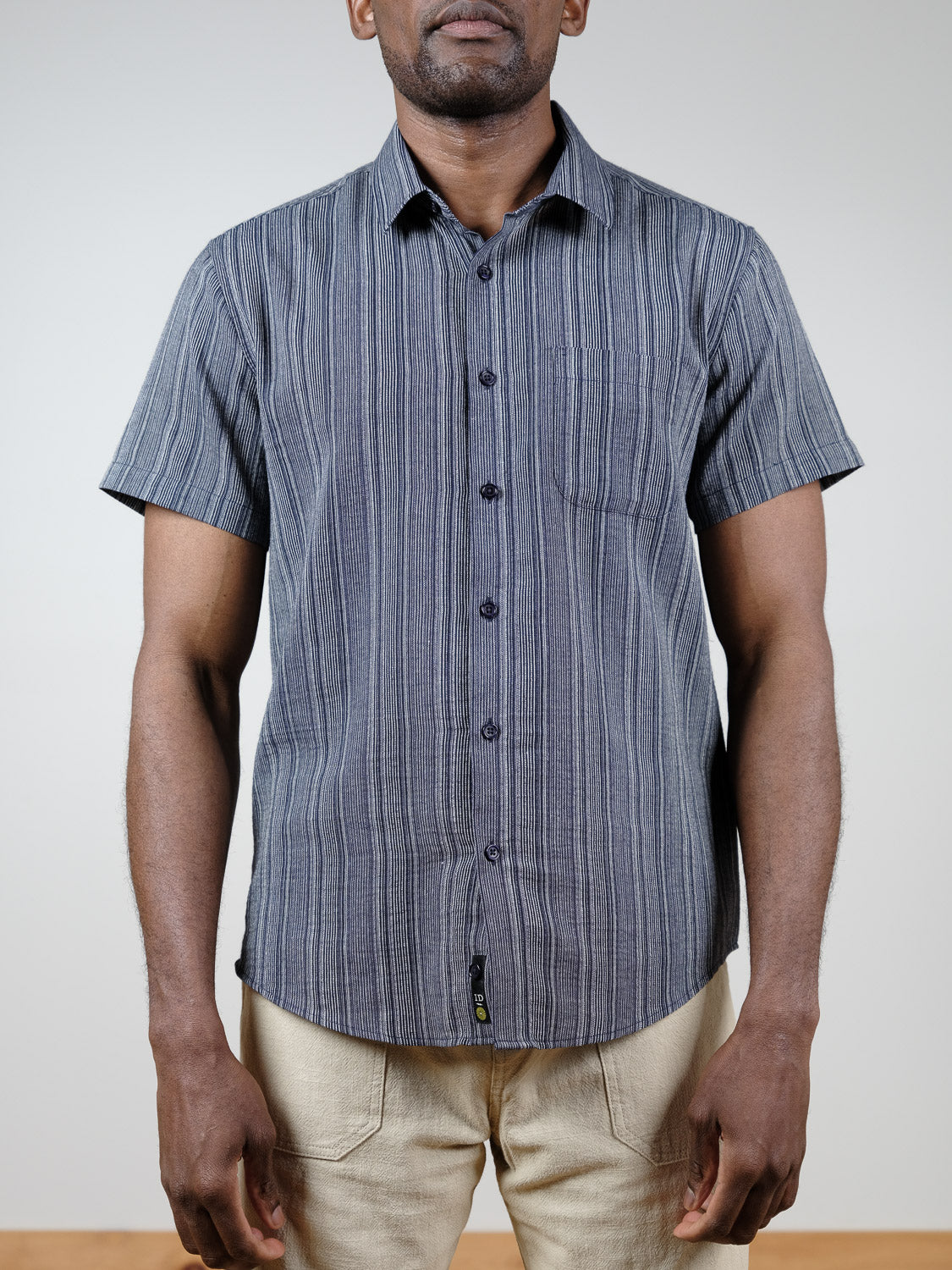 A man in the ID Menswear Strider Short Sleeve Shirt, a relaxed-fit, button-down dark gray striped cotton top, pairs it with beige pants and stands facing forward against a plain light background.