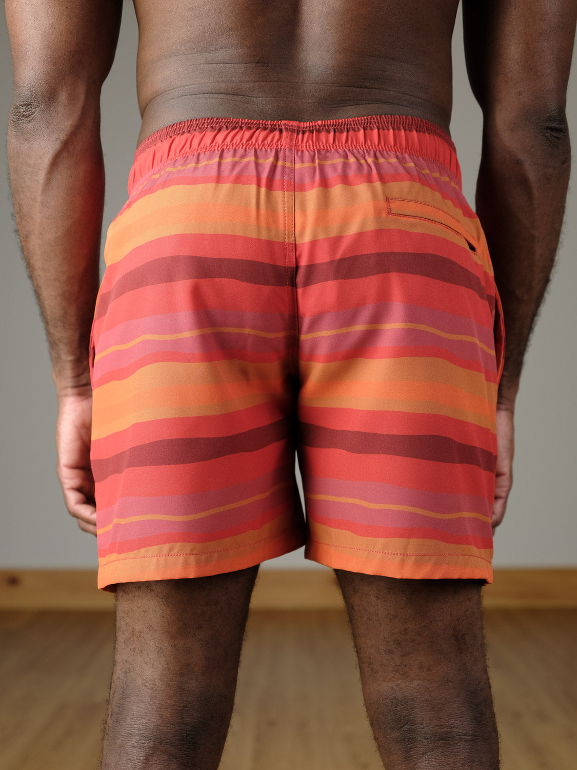 A person stands indoors on a wooden floor, seen from behind, wearing ID Menswear Kochi Fast Dry 4-Way Stretch Swim Trunk in red and orange stripes, featuring an elastic waistband and a back pocket.