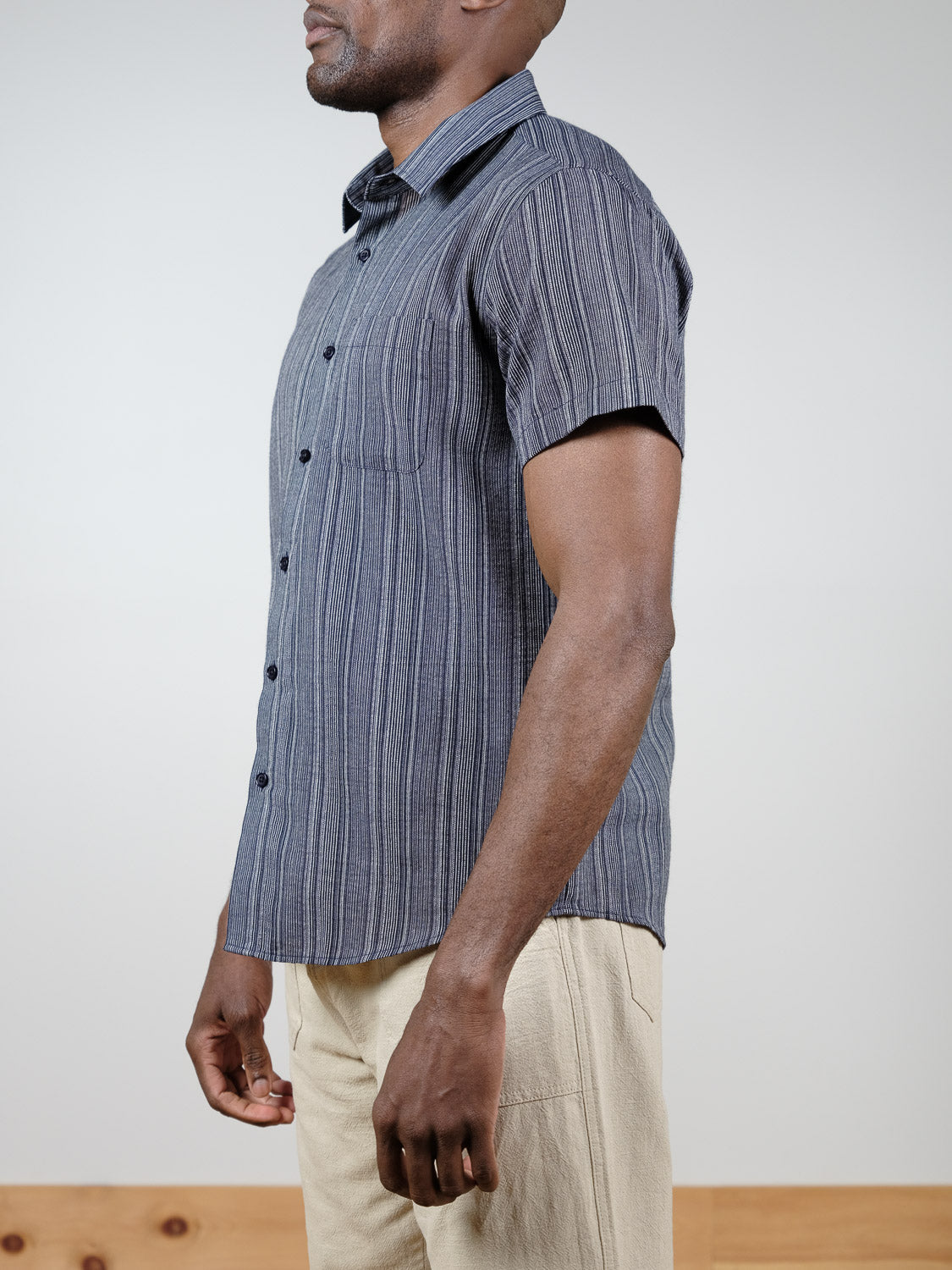 A man stands in profile against a plain background, wearing the ID Menswear Strider Short Sleeve Shirt—a relaxed fit, blue striped cotton shirt—paired with light beige pants for a comfortable, breathable look.