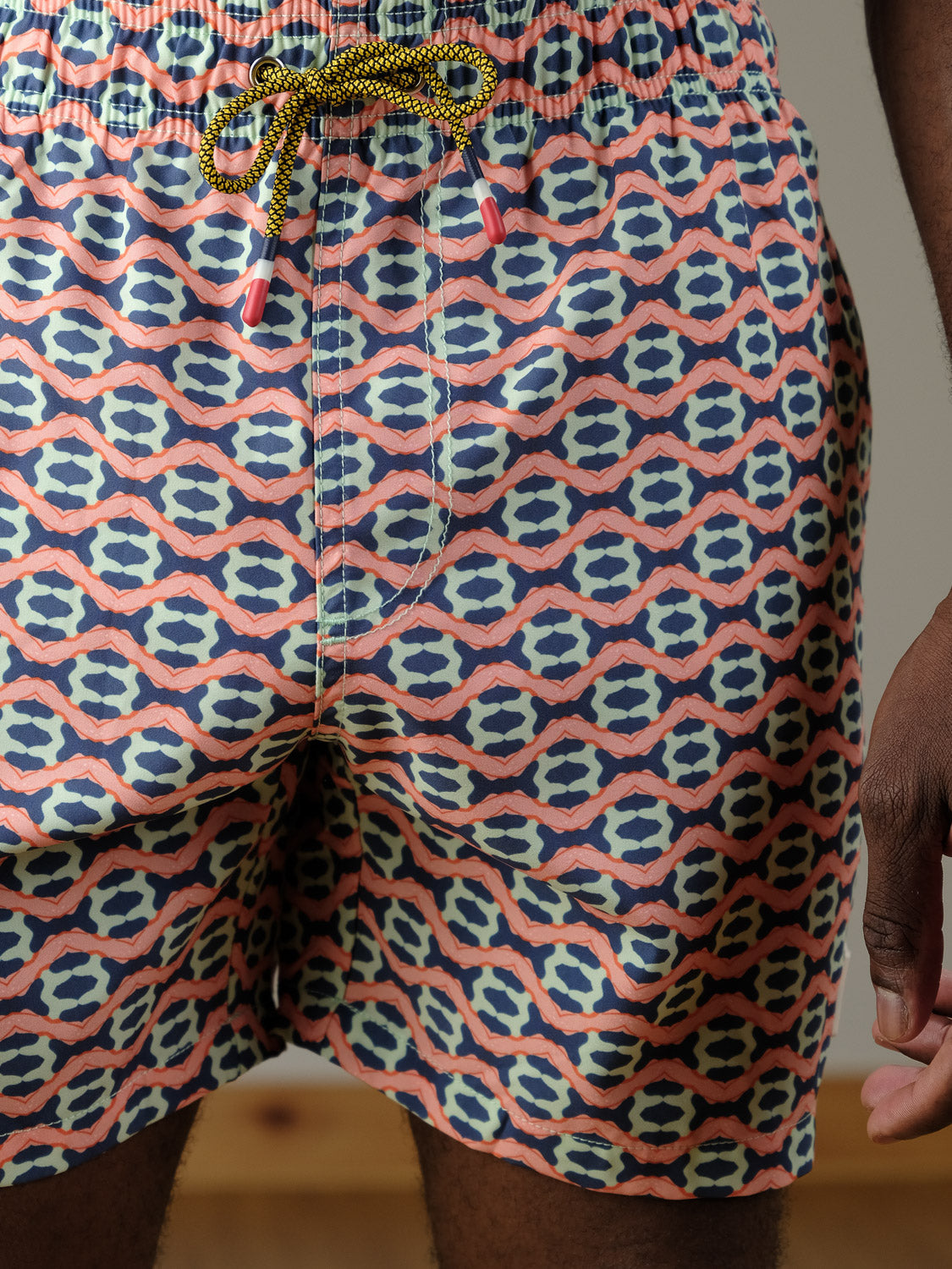 A close-up of a person wearing the Marbella Fast Dry 4-Way Stretch Swim Trunk by ID Menswear, featuring a colorful blue, pink, and white geometric design with a yellow drawstring with red tips. Their right hand rests at their side.